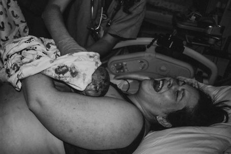 A woman in a hospital bed holds her newborn baby on her chest immediately after birth, expressing intense emotion. A healthcare provider supports the baby, partially covered in a hospital blanket.