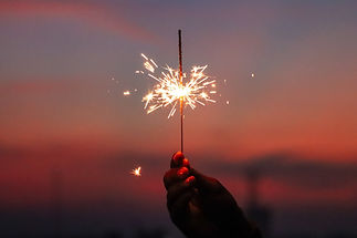 Hand holding a lit sparkler against a dusky sunset background with bright light.