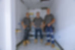 Three male electricians wearing grey company uniforms, tool belts, and work boots, posing with hands on hips inside a small, unfinished room with white walls, exposed wiring, and a blue step ladder behind them.