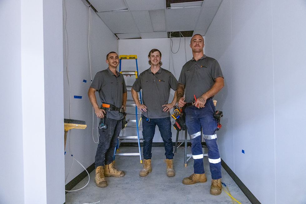 Three male electricians wearing grey company uniforms, tool belts, and work boots, posing with hands on hips inside a small, unfinished room with white walls, exposed wiring, and a blue step ladder behind them.