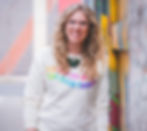 A photo of Jenn Hart with long blond curly hair, a smile and rainbow text shirt that saya focus on all things good