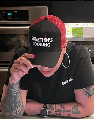 picture of chef with her head tilted down with a hat that says something's cooking