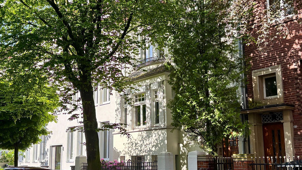 Denkmalgeschützte Mehrfamilienhäuser in einer von Bäumen gesäumten Straße, Autos und Fahrrad im Vordergrund. Sonnige Atmosphäre, lebendige Grüntöne.
