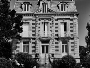 Schwarzweißfoto eines historischen Gebäudes mit Verzierungen, Bäumen im Vordergrund. Das Gebäude hat mehrere Balkone und hohe Fenster.