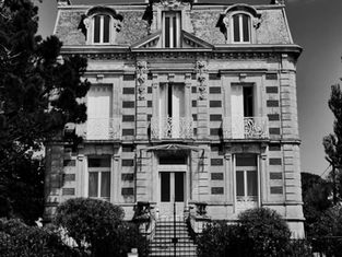 Schwarzweißfoto eines historischen Gebäudes mit Verzierungen, Bäumen im Vordergrund. Das Gebäude hat mehrere Balkone und hohe Fenster.