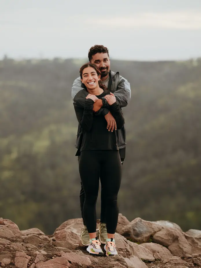 Couple in outdoor athletic clothing hugging on rocky terrain with green mountains in the distance.