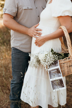 Expecting couple standing close together in a field, holding baby ultrasound photos and a basket of flowers, photographed by Osondu Digital, Central Valley maternity photographer.