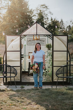 Woman stands in front of a greenhouse holding a bouquet outdoors on the grass.