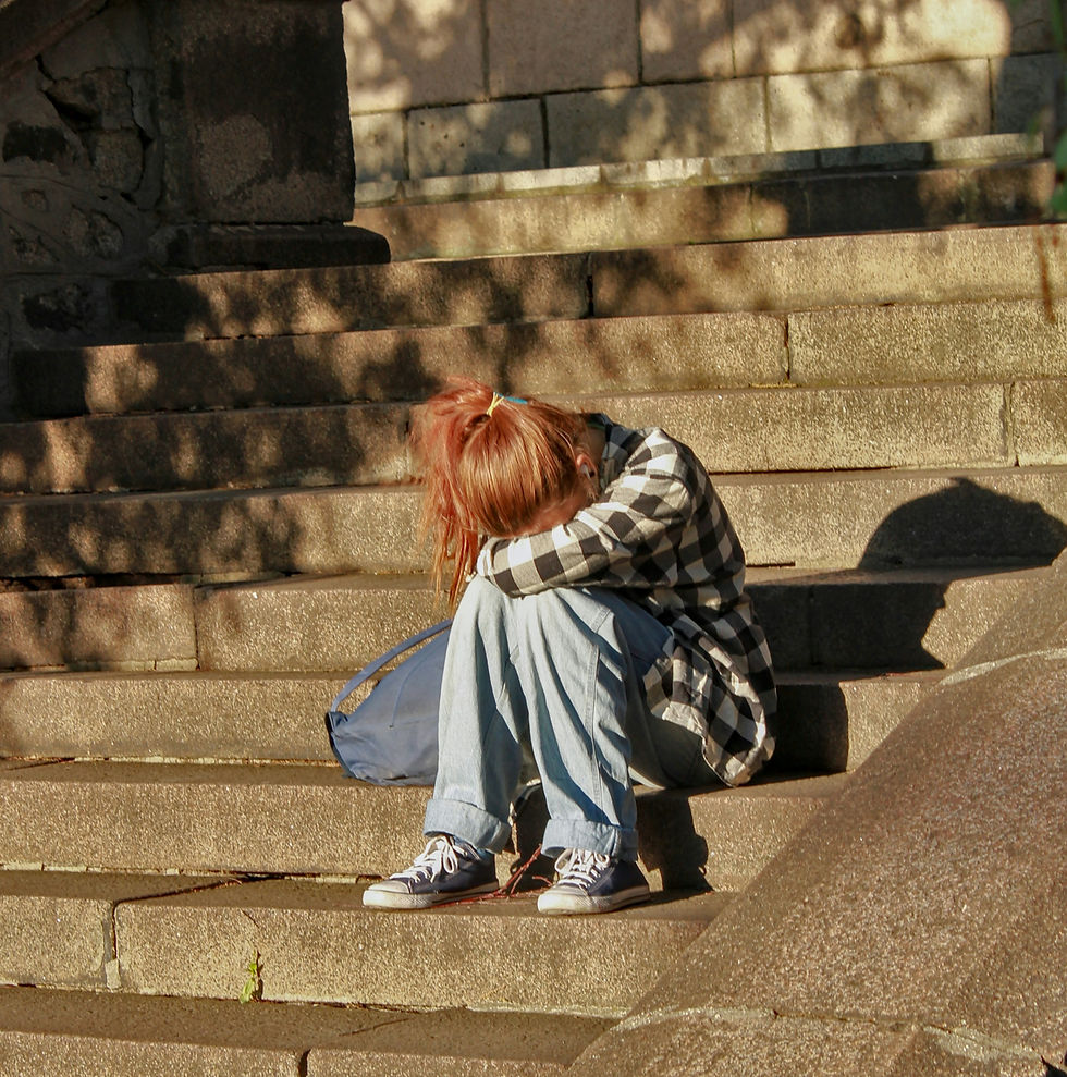Moça sentada em uma escadaria pública com a cabeça baixa apoiada no braço e o braço fechado sob os joelhos