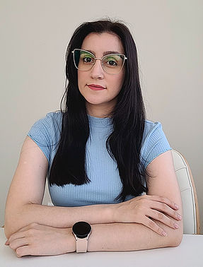 Moça branca de óculos e cabelo médio com tom de castanho mais escurto, usando camiseta azul pastel e está sentada com os braços apoiados sob a mesa e um apoiado sob o outro