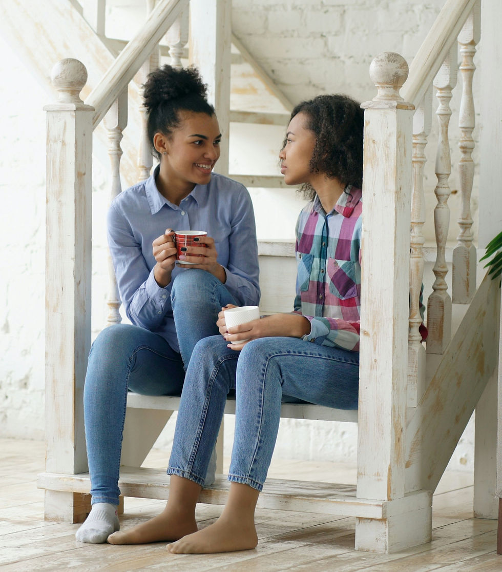 Duas moças sentadas no fim da escada de madeira de uma casa conversando com cada uma com uma caneca na mão