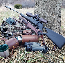 A collection of hunting gear including rifles, scopes, and outdoor equipment laid out on camouflage fabric