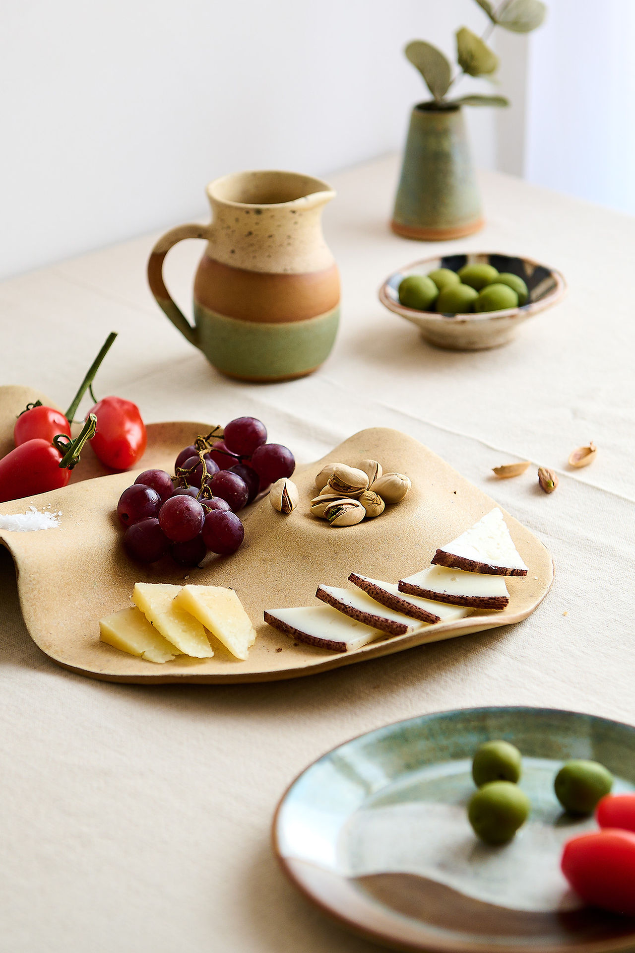 Lifestyle product photography for makers showing a styled scene with handmade ceramics including a charcuterie plate, glazed jug, olives, tomatoes and natural styling details in soft light.