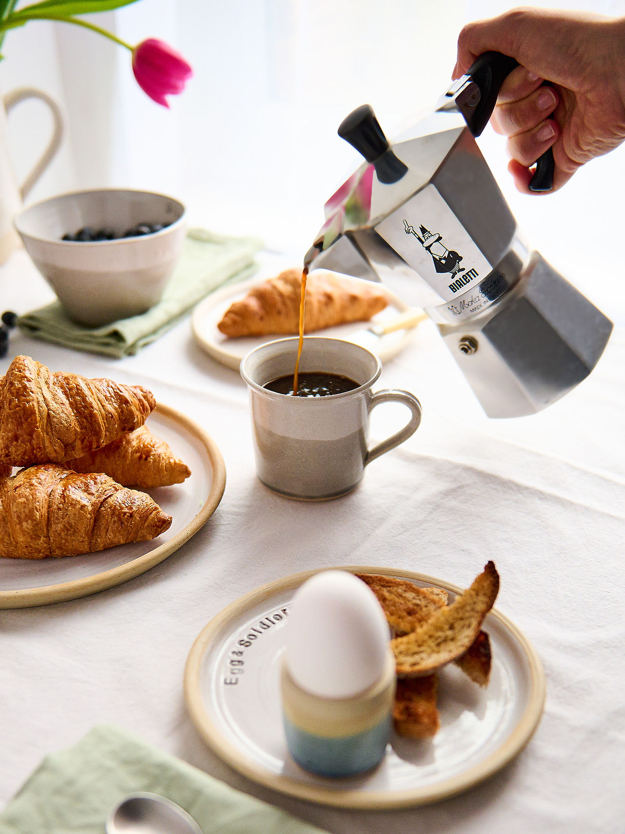 Pouring coffee into a white and grey ceramic mug with breakfast scene of croissants, eggs, and fruit, styled for product photography for handmade ceramics