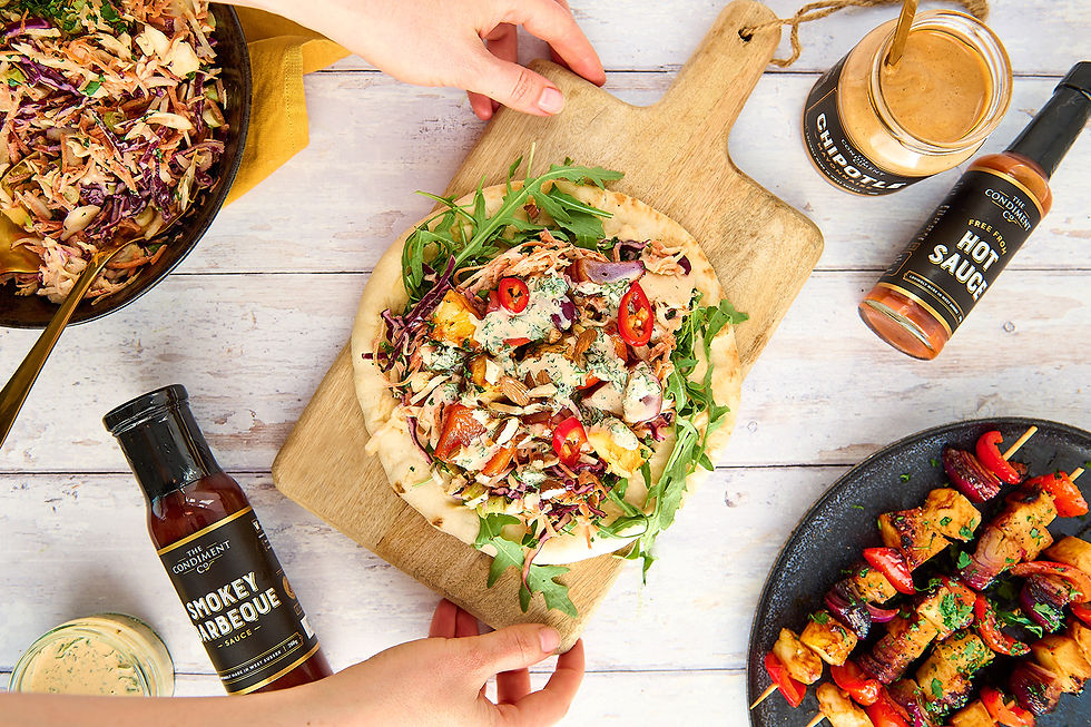 An overhead flatlay from a food content shoot with a flatbread on a wooden chopping board and jars of condiments next to the board. A person's hands are reaching in to pick up the chopping board.