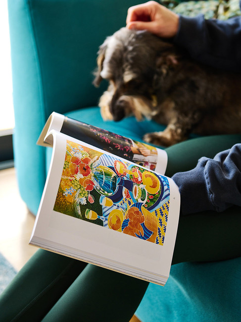 Woman sitting on a teal velvet sofa reading a book about artist Annie Soudain’s life and artwork, with a dog beside her in a cosy lifestyle scene.