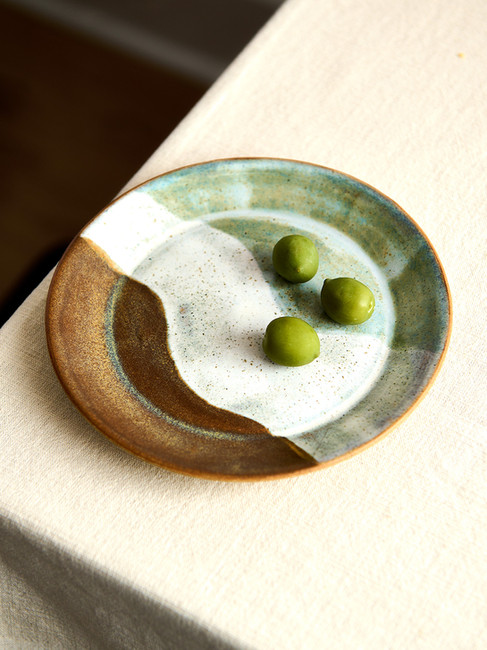 Simple shot of a ceramic side plate on the corner of a table, featuring a three-tone glaze with three olives placed on it.