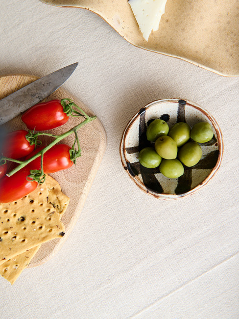 Overhead shot of a small ceramic bowl with green olives, a wooden chopping board, a knife and vine tomatoes styled on a table.