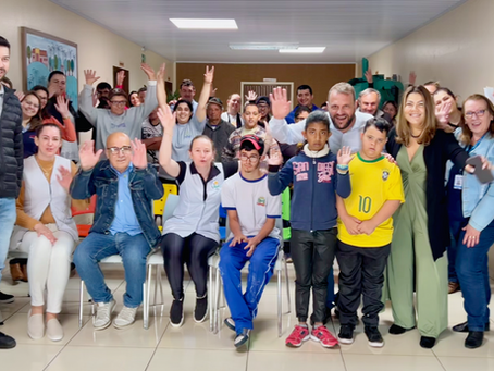 rupo grande de pessoas reunidas em um corredor interno da Apae de Esperança do Sudoeste, no Paraná. Ao centro estão Rogério Carboni, sua esposa Andrea Oliveira, Werther (vice-presidente da FEAPAES), representantes do CEDCA, da gestão da Apae e vários alunos. Todos sorriem, alguns acenam e fazem gestos de inclusão com as mãos. O ambiente é iluminado, com paredes claras e cadeiras coloridas.