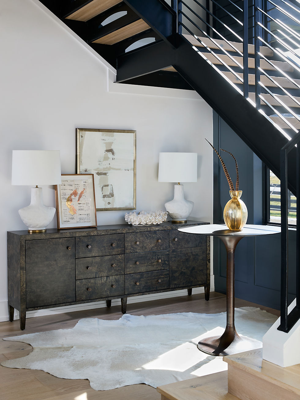 Stylish living area with a dark wooden sideboard, white lamps, abstract art, a round table with a gold vase, and a cowhide rug under stairs.