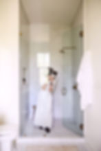 Person in a white dress holds a book in a marble shower. Glass door, towel on railing, light coming through a window. Calm, indoor scene.