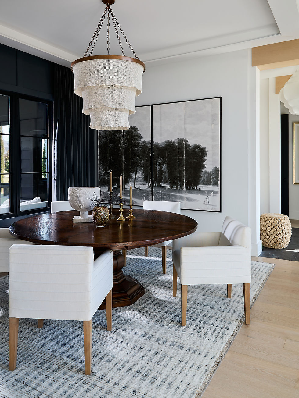 Elegant dining room with a round wooden table, white chairs, and a layered chandelier. Large monochrome art and blue curtains in the background.