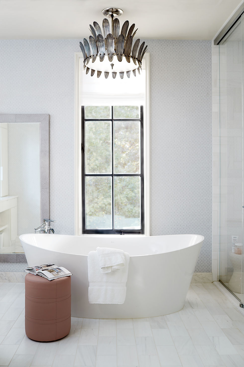 Luxurious bathroom with a white freestanding tub, towel, and open magazine. Elegant chandelier, black window, and beige stool add style.