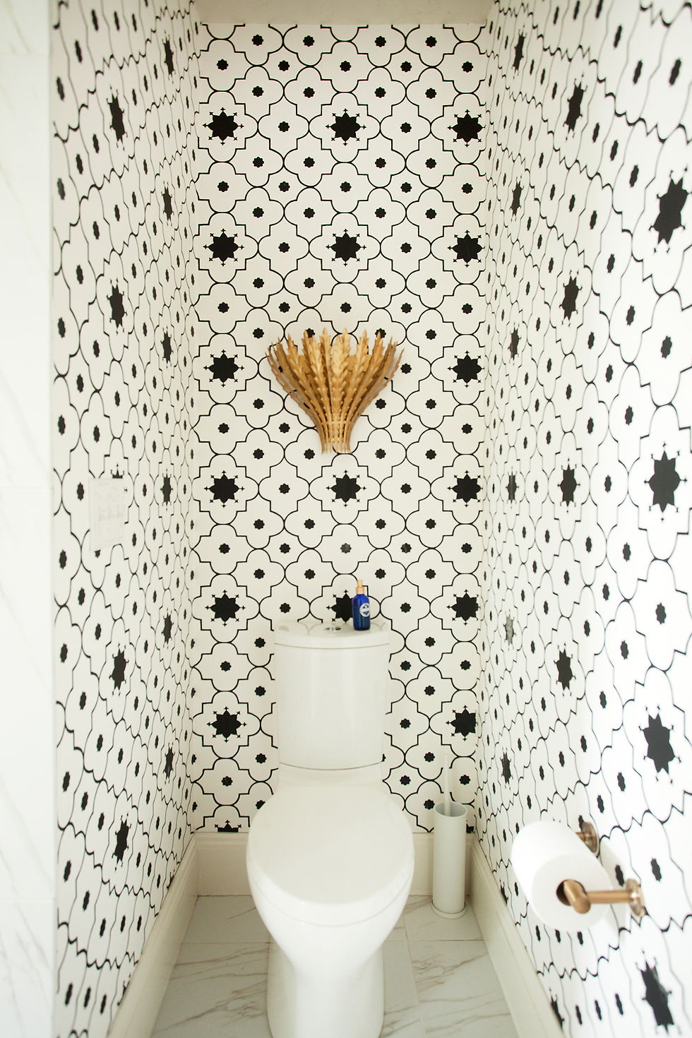 White toilet in a bathroom with black and white patterned wallpaper. A decorative brass sconce t is above the toilet tank. Marble floor.