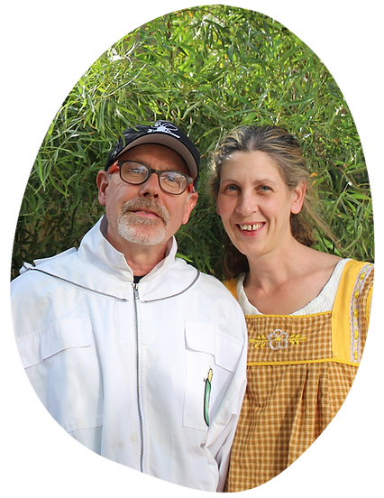 Waist-up outdoor portrait of beekeepers Pete and Betsy, standing side by side in front of lush green foliage. Pete wears glasses, a white beekeeping jacket, and a black cap, and Betsy wears a yellow checkered pinafore over a white blouse, both smiling at the camera.