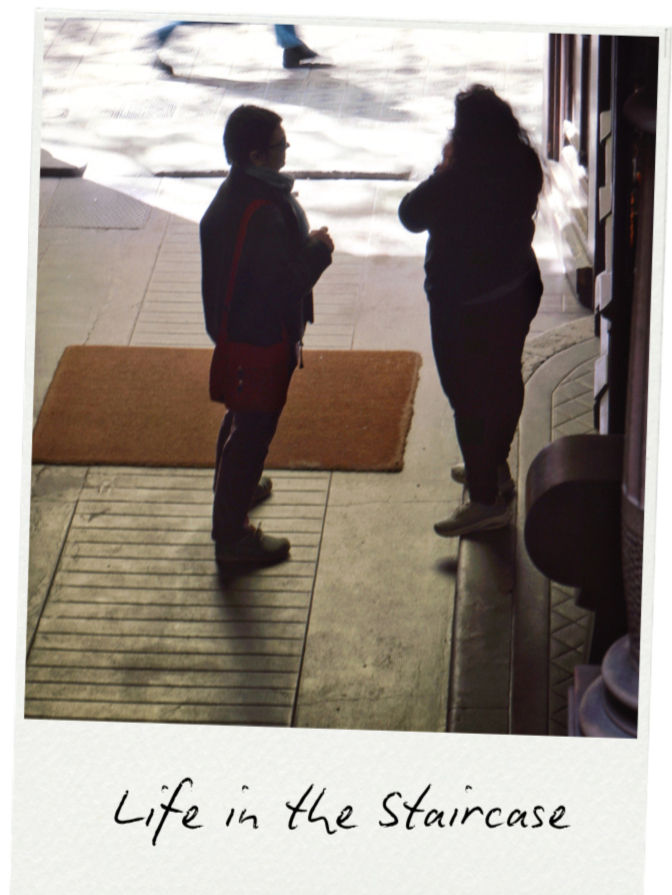 Two people stand and talk in an entryway with a brown mat. Shadows suggest an outdoor scene. Text reads "Life in the Staircase."