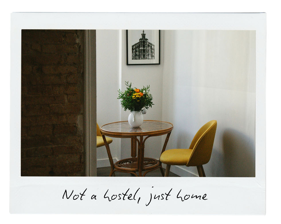 Cozy room with a round table, yellow chair, and vase of flowers. Brick wall on left, framed photo on back wall. Text: "Not a hostel, just home".