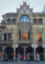 Ornate building facade with intricate carvings and lit windows. People walk by on a cobblestone street.