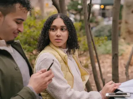A couple sits in a park the man in smiling while on his phone. The woman looks at him with concern. The image illustrates emotional disconnection in a relationship.