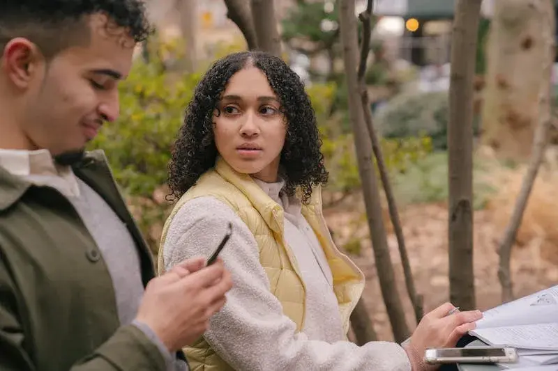A couple sits in a park the man in smiling while on his phone. The woman looks at him with concern. The image illustrates emotional disconnection in a relationship.
