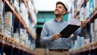 Warehouse manager wearing a hard hat and holding a clipboard while inspecting inventory shelves, representing real-world logistics and supply chain operations in LAPU’s online degree program.