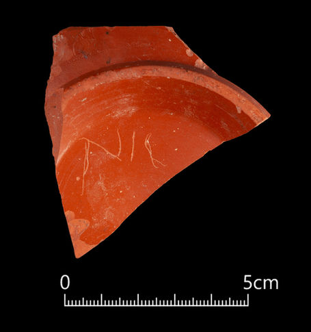 A triangular-shaped sherd of orangey-red pottery on a black background. It is broken off a circular dish, the rest of which is now missing. Three letters have been roughly scratched on the underside, reading the first two of which are legible as N and I.