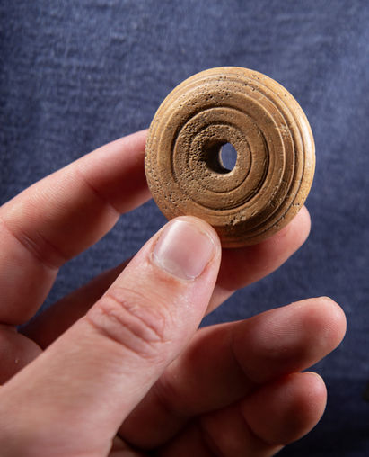 A hand holds up a spindle whorl. The whorl is circular, with a hole in the middle. It is of a light yellowish brown, the colour of antler or bone. There are concentric circles inscribed over the surface.