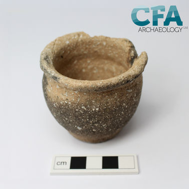 A small ceramic pot sits of a greyish white background. The pot is small, only about five centimetres wide. It is made of light brown clay that has turned black in places around the outside from when it was fired, and has lots of white shell inclusions throughout.