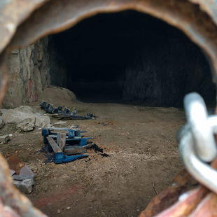 Rusty circular frame views an empty cave with a dirt floor, scattered equipment, and blue pallets. The setting feels abandoned and eerie.