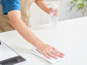 Person wipes a white kitchen counter with a cloth and spray bottle near a stove in a bright, clean kitchen.