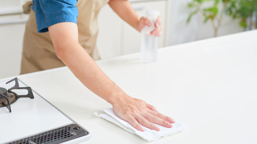 Person wipes a white kitchen counter with a cloth and spray bottle near a stove in a bright, clean kitchen.