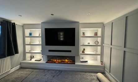 A garage conversion gurus' modern living room featuring built-in shelving with decorations, a mounted television above an electric fireplace, and soft lighting.
