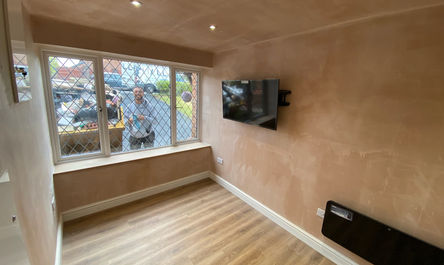 A freshly renovated room with wooden flooring and a wall-mounted TV, adjacent to a residential building, showcases a person visible through the window.