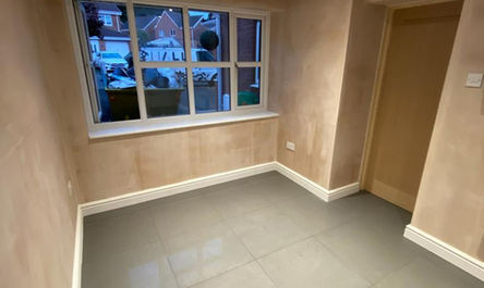 An empty room, previously a garage before its transformation by garage conversion gurus, with beige walls, gray floor tiles, and a window providing a view of the outside area.