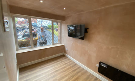 An empty room, transformed from a garage by garage conversion experts, with wooden flooring, a flat-screen TV mounted on the wall, and a window through which a person is visible outside.