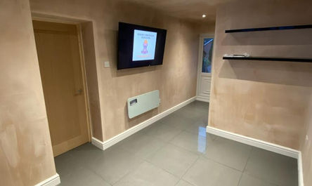 A modern garage conversion in Dukinfield with beige walls featuring a mounted flat-screen tv displaying a graphical icon, floating shelves on the right, and a wooden door on the left.