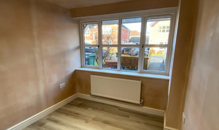 Empty room with a window and a radiator under it, freshly painted walls, and laminate flooring, with a narrow gravel path on its side leading to a garden area.