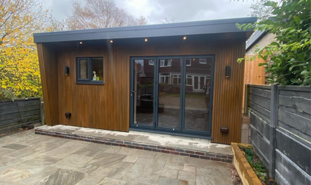 Modern garage conversion office with wooden cladding, sliding glass doors, and an adjacent paved patio area.