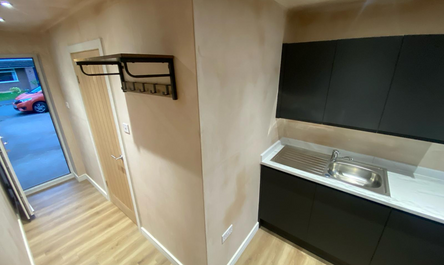 Compact kitchenette with modern black cabinetry and a stainless-steel sink, next to a wall-mounted coat rack in a small room with wooden flooring, showcasing a modern shed with a roller shutter door.