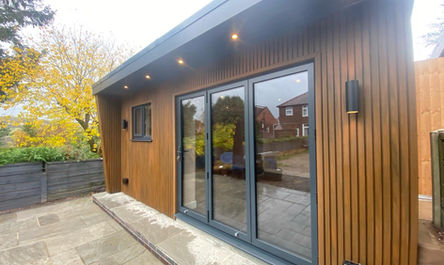 Modern garden room with wooden cladding and large sliding glass doors, surrounded by greenery, featuring outdoor lighting and a paved patio area.